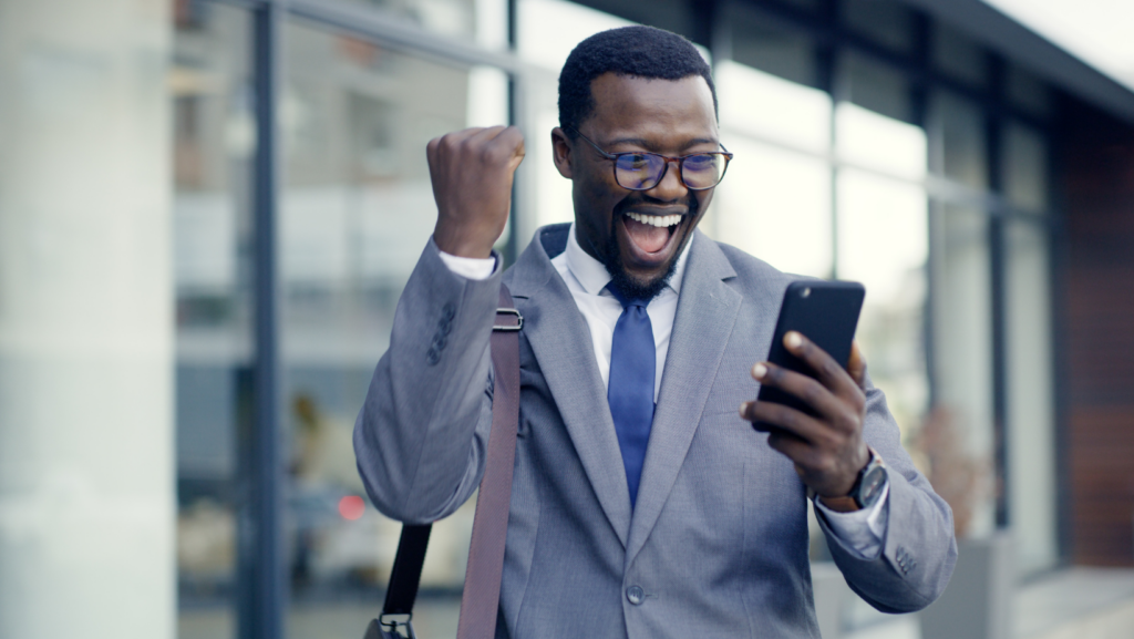 A professionally dressed person in a gray suit stands outdoors near a modern glass building, holding a smartphone and raising a fist in an enthusiastic, celebratory gesture.