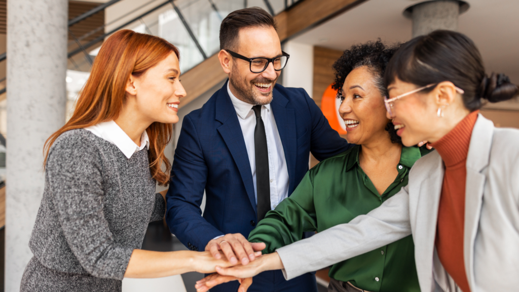 A group of four coworkers standing in a modern office space, placing their hands together in a collaborative team huddle.