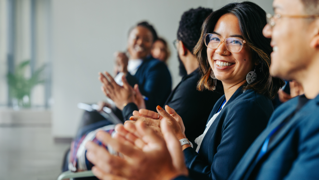 A group of people seated in a row during a professional meeting or presentation, with several individuals clapping. In the foreground, a person wearing glasses and a dark blazer looks toward the camera while applauding, with others slightly out of focus nearby, also clapping. The setting appears to be a bright indoor conference or office space with soft natural lighting and a blurred background.