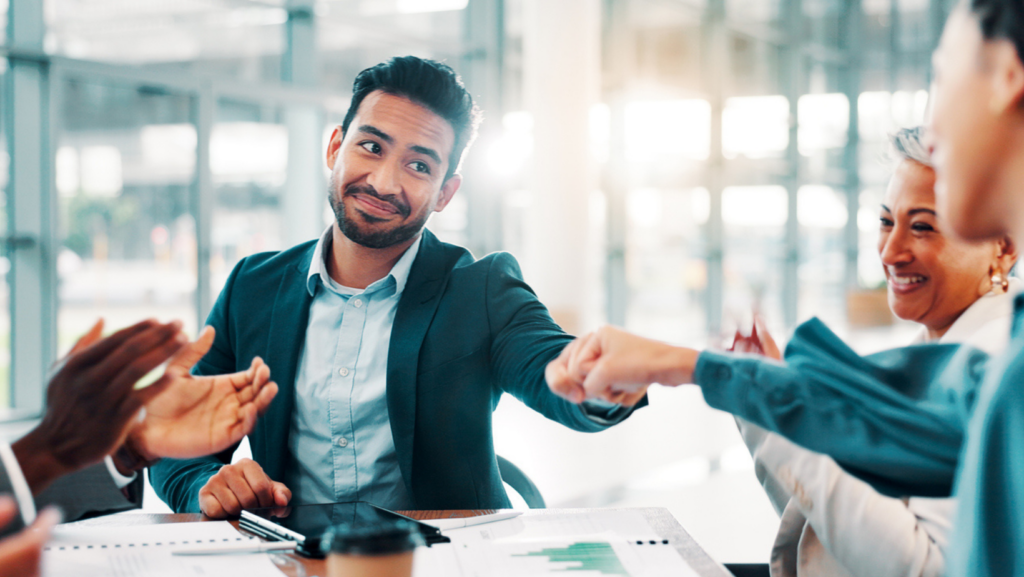 A group of coworkers sit around a conference table, smiling as a man in a suit fist-bumps a colleague during a meeting. Papers, charts, and a coffee cup are on the table in front of them.