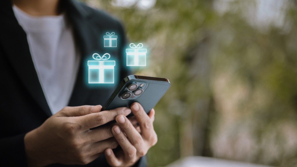 Close-up of a person holding a smartphone outdoors, with three glowing digital gift box icons floating above the screen, suggesting mobile shopping, rewards, or gifting features.