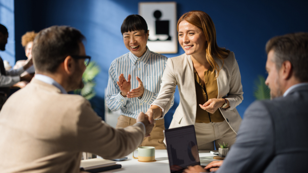 Group of four professionals gathered around a table in a modern office, with one person standing and shaking hands with a seated colleague while another stands nearby clapping; a laptop, coffee mug, and small plant sit on the table against a blue wall with framed artwork.
