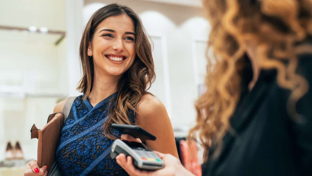 Person in a sleeveless blue lace top holding a brown clutch and shoulder bag while making a contactless payment at a retail checkout. A cashier holds a card reader and smartphone in the foreground, with a softly blurred store interior in the background.
